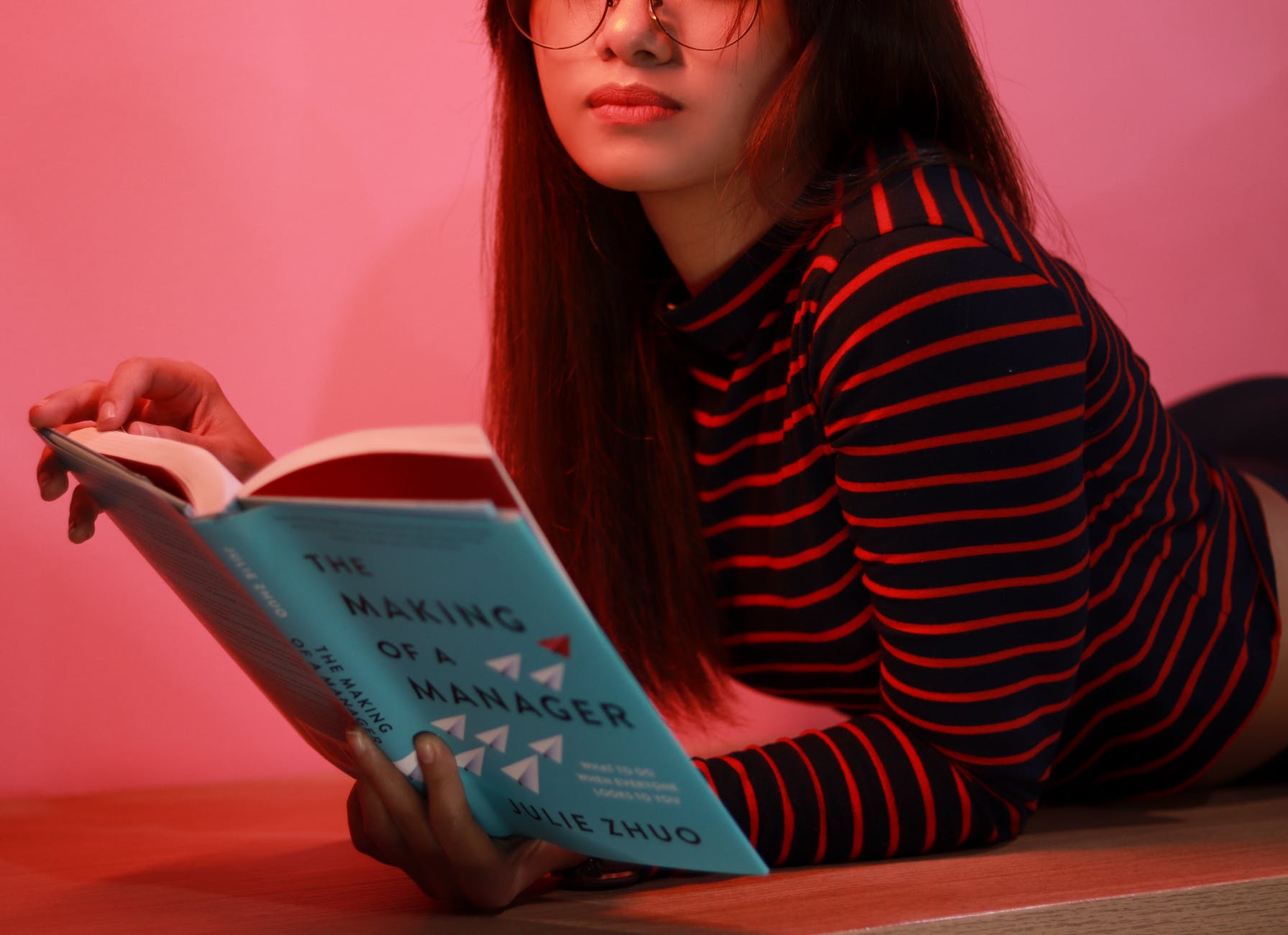 Una chica pelirroja tumbada boca abajo leyendo un libro. Usa gafas y viste una camiseta manga larga de color negro y franjas rojas. 
